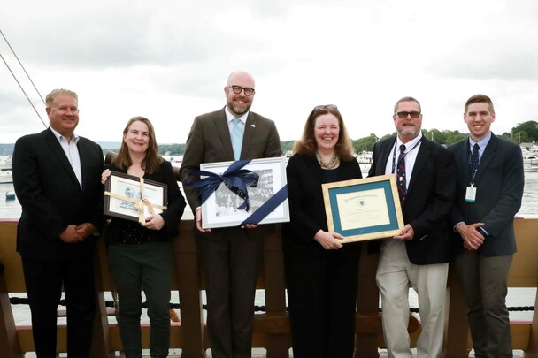 Dignitaries and Museum leadership stand aboard Mayflower II holiding commemorative objects.