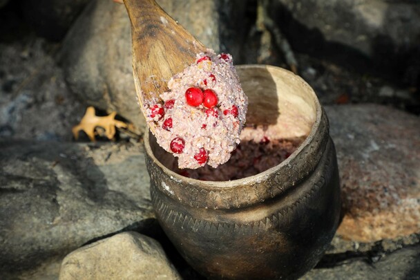 Nasaump with cranberries cooks over an outdoor fire. A Museum educator lifts some of the mixture on a wooden spoon.