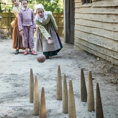Pilgrim children play a bowling game near a grey, clapboard house.