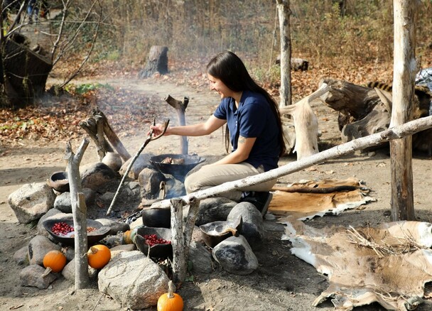 Educator cooks over a fire at the Historic Patuxet Homesite. Red cranberries sit in wooden bowls on rocks around the fire along with orange pumpkins.