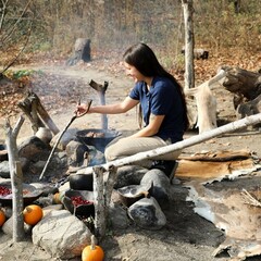Educator cooks over a fire at the Historic Patuxet Homesite. Red cranberries sit in wooden bowls on rocks around the fire along with orange pumpkins.