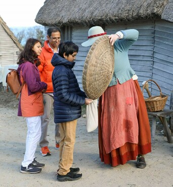 A family of three examines a basket held up by a Pilgrim woman in the 17th-Century English Village.