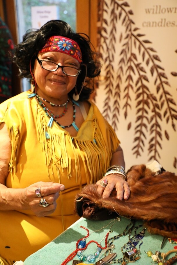 A women dressed in Wampanoag regalia sits at a table with jewelry and fur items that she has made.