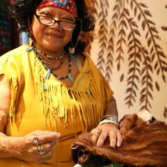A women dressed in Wampanoag regalia sits at a table with jewelry and fur items that she has made.