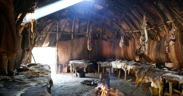 Furs line the interior of a wetu. A fire is going in the center of the structure and sunlight beams in.