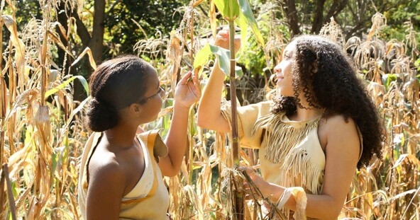 Two young Wampanoag women dressed in regalia check on growing corn at the Historic Patuxet Homesite.