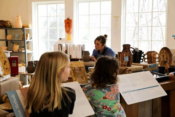 An artisan works to create pottery in the Craft center while two young guests observe her.