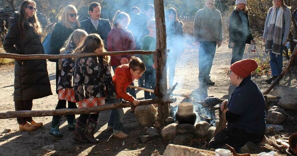 Children engage with a Museum Educator at the Historic Patuxet Homesite Cooking Arbor. Adults stand on the site and observe.