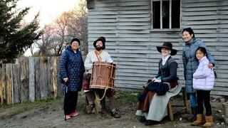 Two women and young girl pose with seated Pilgrims in front of a grey Pilgrim house.