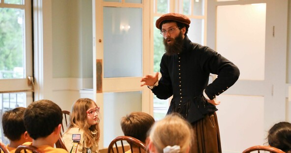 An educator dressed in Pilgrim clothing speaks to a group of seated, young scouts.