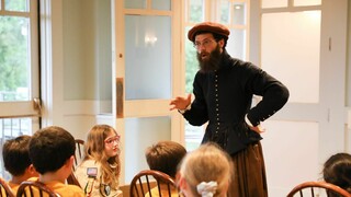 An educator dressed in Pilgrim clothing speaks to a group of seated, young scouts.