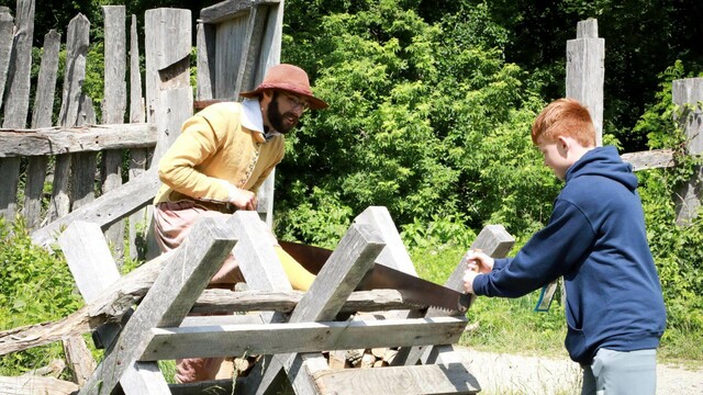 A modern schoolchild helps a Pilgrim saw wood