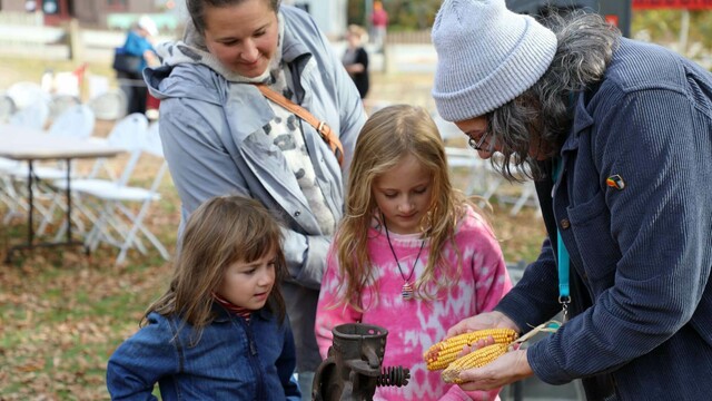 Plimoth Grist Mill Educator shows dried corn to two young girls as their mother watches.