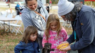 Plimoth Grist Mill Educator shows dried corn to two young girls as their mother watches.