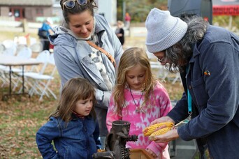 Plimoth Grist Mill Educator shows dried corn to two young girls as their mother watches.