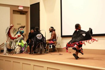 Members of Standing Quiver sit around a drum while a young child demonstrates a dance.