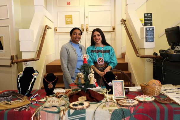 Two Indigenous Artisan Fellows stand behind a table displaying their art.