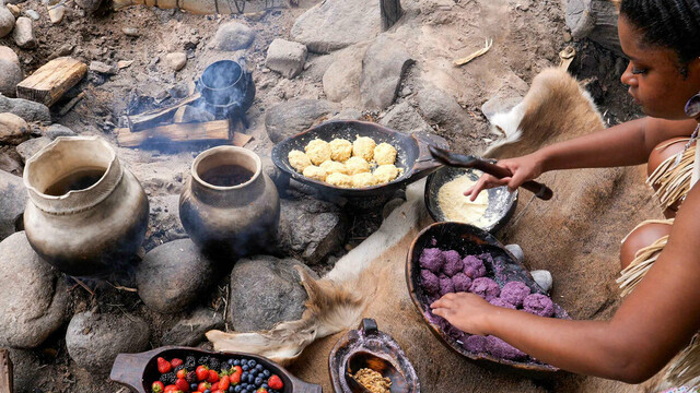Educator in Wampanoag regalia cooks corn cakes and roasts fish over a fire at the Historic Patuxet Homesite.