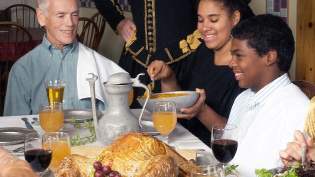 Guests seated at a table enjoy thanksgiving food. A Pilgrim stands behind them.