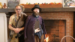 Indigenous Educator dressed in regalia and Educator dressed as a Pilgrim stand in front of a brick fireplace.
