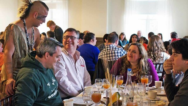 Indigenous Educator dressed in regalia speaks with guests seated for Thanksgiving dinner.
