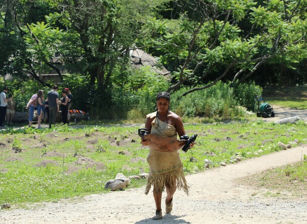 Educator in Wampanoag regalia carries a large wooden bowl filled with quahogs across the Historic Patuxet Homesite on a summer day
