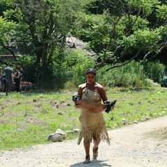 Educator in Wampanoag regalia carries a large wooden bowl filled with quahogs across the Historic Patuxet Homesite on a summer day