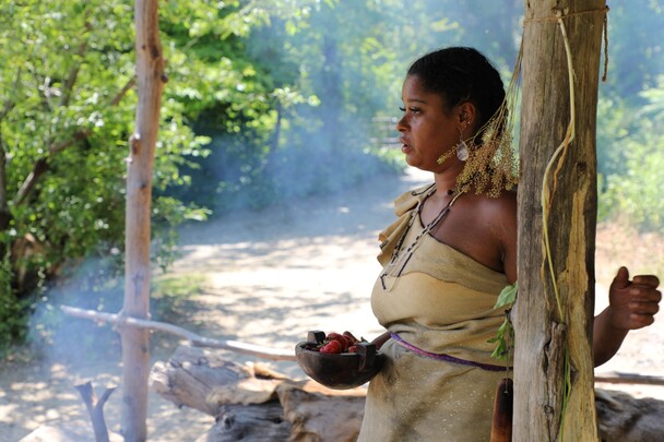 Indigenous Educator dressed in Wampanoag regalia holds a wooden b owl of strawberries as she stands under the Cooking Arbor on the Historic Patuxet Homesite