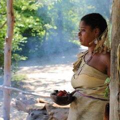 Indigenous Educator dressed in Wampanoag regalia holds a wooden b owl of strawberries as she stands under the Cooking Arbor on the Historic Patuxet Homesite