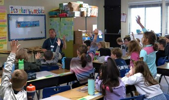 A Museum Educator visits an elementary school classroom. Children raise their hands to ask questions.