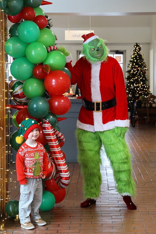 Grinch poses with a young Museum guest in front of a green and red balloon arch.