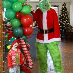Grinch poses with a young Museum guest in front of a green and red balloon arch.
