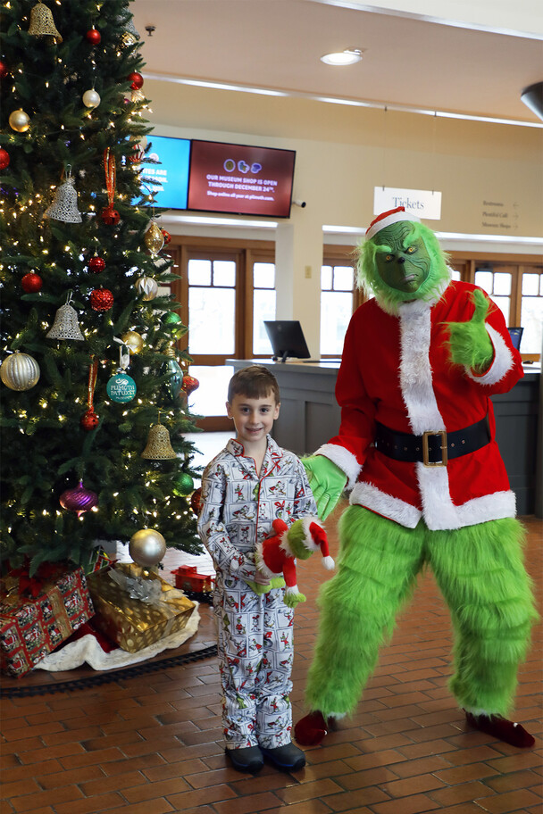 Grinch poses with boy attending the matinee dressed in pajamas in front of a decorated Christmas tree.