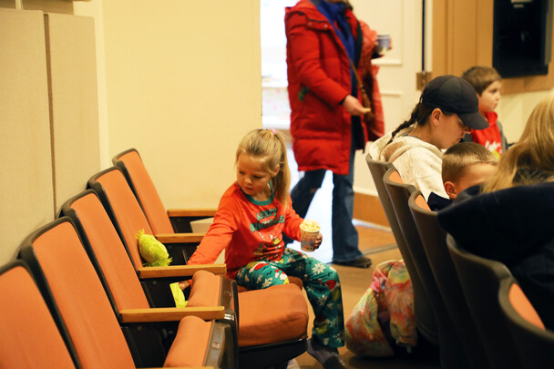 Child in pajamas sets a stuffed animals of the Grinch into a movie theater seat.