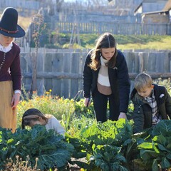 Three children help a Pilgrim woman garden in the English Village.