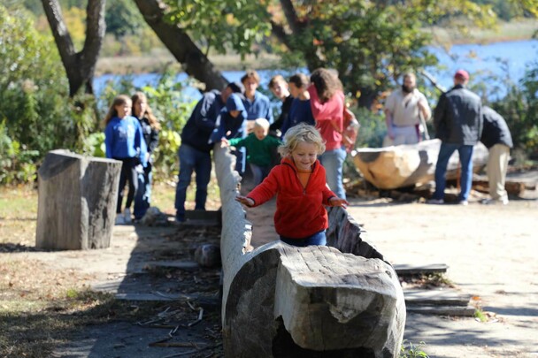 A young, blonde boy in a red jacket runs in a large mishoon. Children behind him enjoy the Historic Patuxet Homesite.