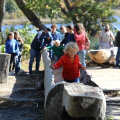 A young, blonde boy in a red jacket runs in a large mishoon. Children behind him enjoy the Historic Patuxet Homesite.
