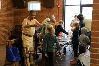 Museum Educator holds a wampum necklace before young learners. Children and adults are gathered at a craft table behind them.