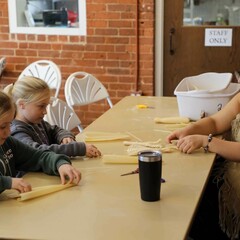 Museum Educator, dressed in Wampanoag regalia, instructs two young, blonde girls, in making cornhusk dolls.
