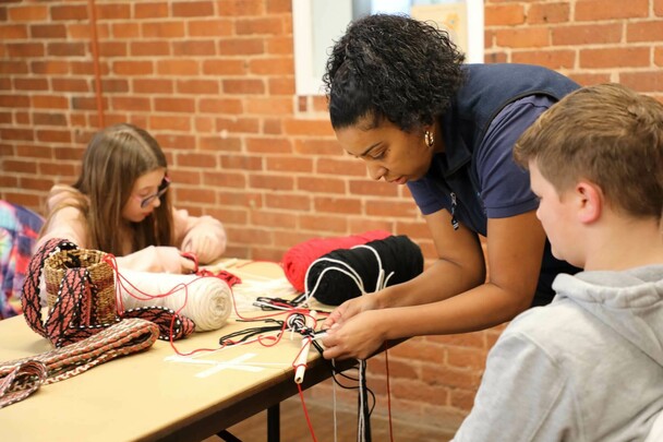 Museum Educator assists a boy with his finger weaving project. A young girl makes her own creation across the table from them.