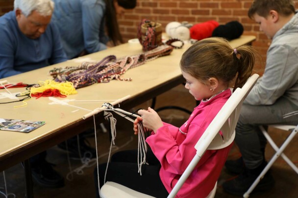 A young girl practices finger weaving at a table with people of all ages who are practicing the craft as well.