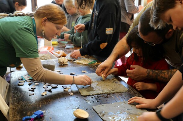 Educators and young learners are gathered around a table, making crafts with slabs of wet, grey clay and white and purple wampum.