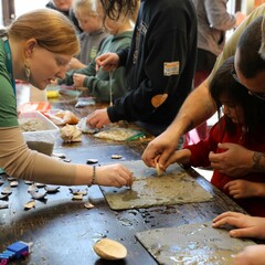 Educators and young learners are gathered around a table, making crafts with slabs of wet, grey clay and white and purple wampum.