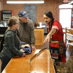 Educator points to an archery bow placed on a wooden table and speaks with an man and woman.