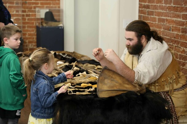 Museum Educator, seated at a table with cultural objects, engages with a young boy and girl who examine the items on the table.
