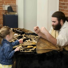 Museum Educator, seated at a table with cultural objects, engages with a young boy and girl who examine the items on the table.