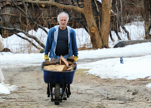 Museum staff moves a wheel barrel filled with wood across the Historic Patuxet Homesite. Snow covers the grounds and the brown winter wetu is in the background.