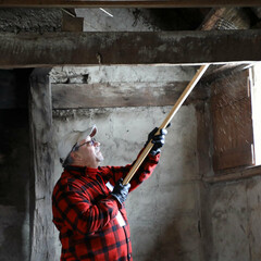 A volunteer dressed in a red flannel shirt raises a wooden broom to sweep above a window inside a Pilgrim house.
