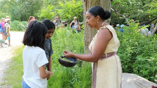 Museum educator in Wampanoag regalia holds a wooden bowl. She lifts a green plant from the bowl to show to two young girls visiting the Homesite.