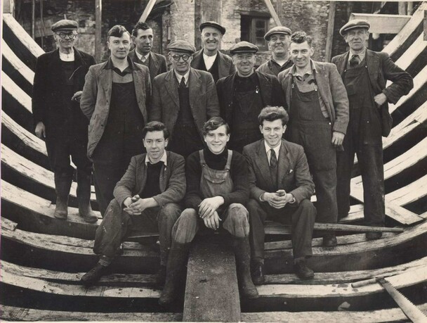 Men pose within partially constructed boat hull